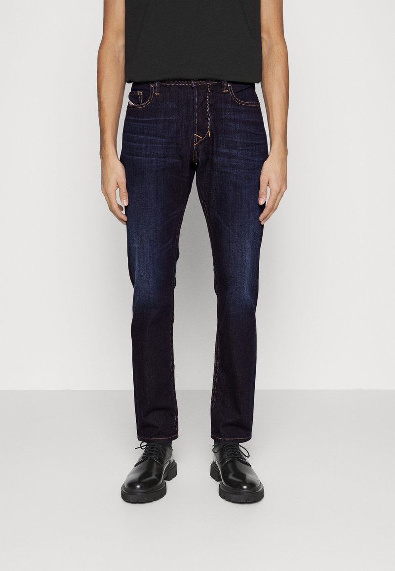 Vaqueros de mezclilla azul oscuro con corte ajustado, presentan costuras naranjas en contraste y cinco bolsillos, combinados con zapatos negros con cordones.