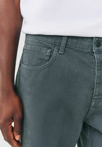 Šedé džíny z denimu s texturovaným povrchem, které mají přední kapsu, poutka na opasek a knoflíkové zapínání. Detailní pohled na látku a design.