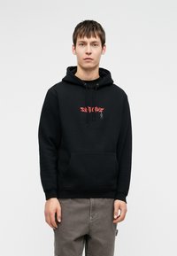 Μαύρη μπλούζα με κουκούλα και μπροστινή τσέπη, με κόκκινο γραφικό κείμενο στο στήθος. Το υλικό φαίνεται μαλακό με άνετη εφαρμογή, φορεμένη με γκρι παντελόνι.