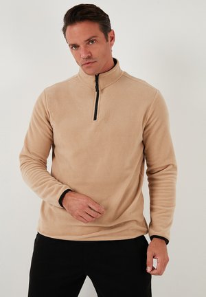 Tan fleece pullover med høj krave og en sort lynlås. Har lange ærmer med sort kant ved manchetten. Glat tekstur.