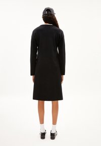 Robe noire à manches longues, coupe droite, longue jusqu'au milieu du mollet, associée à des chaussettes blanches et des chaussures noires ; design minimaliste.