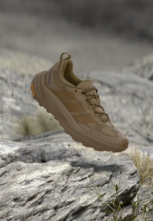 Hellbrauner Adidas Terrex Gore-Tex Traillaufschuh, der über felsigem Gelände mit kleinen Pflanzen und Erdpartikeln schwebt.