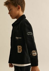 Must mustaga, millel on tikitud Batmani logo, "B" patch ja logo detailid. Omab vabalt liikuvat kraed ja rinna taskut.