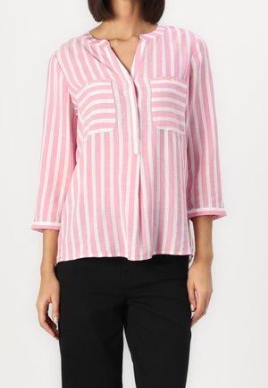Vrouw draagt een roze-wit verticale gestreepte blouse met twee voorzakken en driekwart mouwen, gecombineerd met zwarte broek.