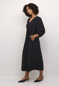 Robe noire à manches longues, avec fermeture à boutons, ceinture à la taille, poches latérales et jupe fluide ; tissu texturé, coupe décontractée et décolleté en V.