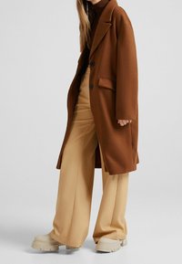 Brązowy wełniany płaszcz o luźnym kroju, z dużymi klapami, zapinany na pojedynczy guzik, zestawiony z beżowymi szerokimi spodniami i masywnymi kremowymi butami.