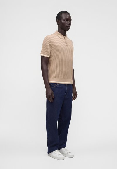 Homme debout portant un polo en maille beige à manches courtes, un jean bleu foncé et des baskets blanches sur un fond clair uni.