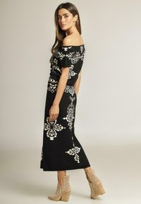 Vestido negro off-the-shoulder con patrones florales blancos, de longitud midi, complementado con botas de tobillo perforadas en tono marrón con tacón apilado.