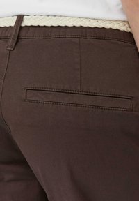 Pantalons en tissu marron avec une ceinture tressée crème et une poche arrière avec un rabat rectangulaire. Le matériau a une texture lisse.