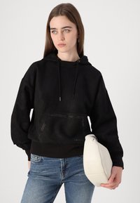 Felpa nera con cappuccio e tasche frontali, caratterizzata da una sottile cucitura a contrasto. Jeans in denim blu chiaro e una borsa a tracolla beige con zip.
