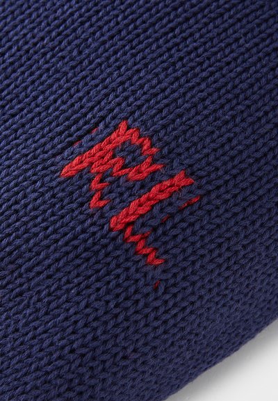 Strikket marineblåt stof med en tekstureret overflade, der har et fremtrædende rødt zigzag broderidesign. Syningen er detaljeret og dristig.