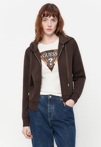 Jonge vrouw met rood haar, gekleed in een bruine zip-up hoodie over een witte Guess logo t-shirt en blauwe jeans, staande tegen een witte achtergrond.