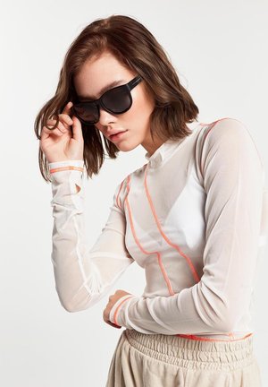 White mesh long-sleeve top with orange geometric patterns, paired with dark sunglasses. Features a high collar and fitted sleeves.