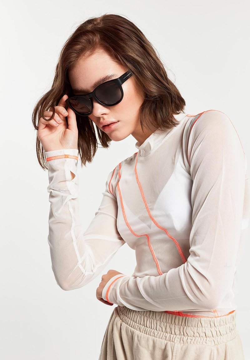 Top lung din plasă alb cu mâneci lungi, cu modele geometrice portocalii, asortat cu ochelari de soare închiși la culoare. Prezintă un guler înalt și mâneci potrivite.