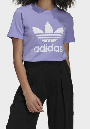 Nainen, jolla on päällään liilanvärinen Adidas-paita valkoisella logolla ja tekstillä, yhdistettynä korkeavyötäröisiin mustiin laskostettuihin housuihin, seisomassa näkyvällä yhdellä kädellä.