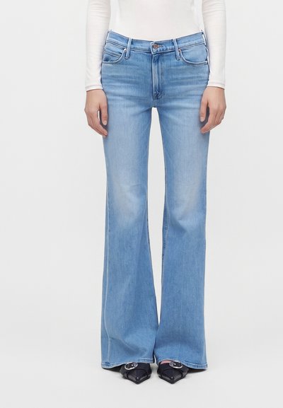 Vue de face d'une personne portant un jean évasé bleu clair, un haut blanc à manches longues et des chaussures noires pointues avec des boucles argentées.
