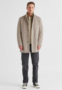 Beiger Wollmantel mit geradem Schnitt, fünf Knöpfen und hohem Kragen, über einem grünen Pullover getragen und kombiniert mit dunklen Jeans und Turnschuhen.