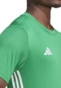 Maglietta sportiva verde realizzata in tessuto traspirante, con scollo rotondo, dettagli in rete e logo Adidas bianco sul davanti.
