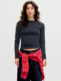 Ung kvinne med bølgete mørkt hår iført en svart langermet skjorte, mørke bukser og en rød rutete skjorte knyttet rundt midjen.