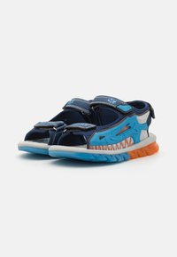 Blå og marineblå sandaler med hai-design, borrelåser, åpen tå og en oransje og blå teksturert såle for godt grep.