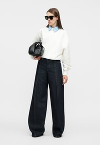 Γυναίκα που φοράει υπερμεγέθη λευκό φούτερ, μπλε πουκάμισο με γιακά, φαρδιά σκούρα τζιν, μαύρα γυαλιά ηλίου, κρατώντας μαύρη δερμάτινη τσάντα.