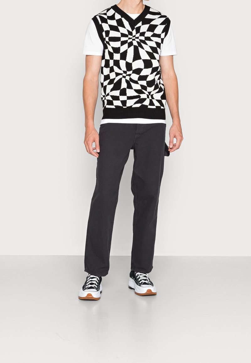 Homme portant un gilet en tricot à damier tourbillonnant noir et blanc, un t-shirt blanc, un pantalon noir et des baskets noir et blanc, debout contre un mur blanc.