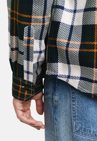 Zielona, biała i granatowa koszula flanelowa w kratę z pomarańczowymi akcentami; posiada mankiety na guziki i teksturę w warkocz. Nosi ją z niebieskimi dżinsami.