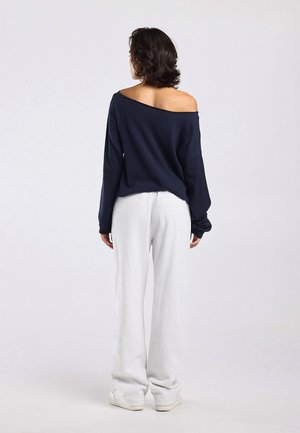 Vrouw staand met de rug naar voren, draagt een marineblauwe off-shoulder top met lange mouwen, lichtgrijze losse broek en witte sneakers op een effen achtergrond.
