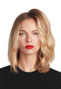 Een vrouw met gelaagd, golvend blond haar die een zwart shirt draagt. Ze heeft helderrode lippen en groene ogen met subtiele eyeliner.