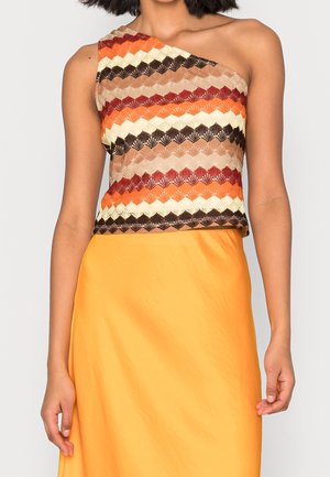 Kvinde iført en skulderstrikket top med zigzag-mønster i brun, orange, creme og rød, kombineret med et højtsiddende, knaldgult skørt.