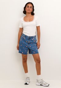 Camiseta blanca de manga corta con detalle de encaje, combinada con shorts de mezclilla azul con bordado floral y un ajuste relajado. Zapatillas de suela de goma y calcetines.