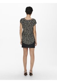 Femme se tenant de dos face à l'appareil photo, portant un haut noir et beige à motifs, une mini jupe noire et des sandales à talons hauts noires avec des brides autour des chevilles.