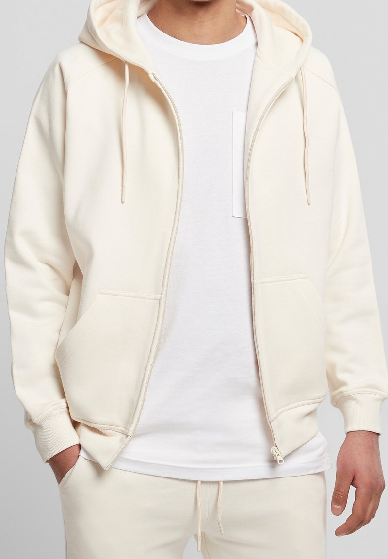 Sudadera con cremallera de color crema en suave tejido de algodón, con capucha, bolsillos frontales y puños ajustados. Lleva puesta una camiseta blanca debajo.