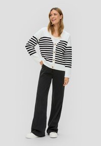 Gestreifter Cardigan in Schwarz-Weiß mit V-Ausschnitt und Knopfleiste, kombiniert mit hochgeschnittenen schwarzen weit geschnittenen Hosen und weißen Sneakers.