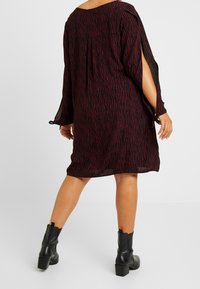 Schwarz-burgunderfarbenes Kleid mit Muster, lockerem Sitz, langen Ärmeln, seitlichen Schlitzen und einem Rundhalsausschnitt. Kombiniert mit knielangen schwarzen Stiefeln.