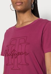 T-shirt di cotone bordeaux con un grande logo "Hilfiger" con accenti ricamati. Design a girocollo con maniche corte. Texture liscia.