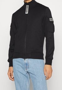 Chaqueta bomber negra hecha de tejido suave, con un cuello alto, cierre de cremallera, parche en la manga y bolsillos laterales. Corte casual.