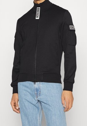 Giacca bomber nera realizzata in tessuto morbido, con colletto alto, chiusura con zip, toppa sulla manica e tasche laterali. Vestibilità casual.