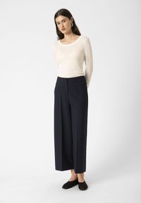 Top a maniche lunghe color crema in un tessuto morbido e liscio, abbinato a pantaloni blu navy a gamba larga realizzati in un materiale strutturato. Scarpe nere slip-on.