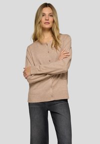 Beige Strickjacke aus weichem Material, mit rundem Ausschnitt, gerippten Bündchen und sechs Knöpfen vorne, kombiniert mit dunkler Jeans.