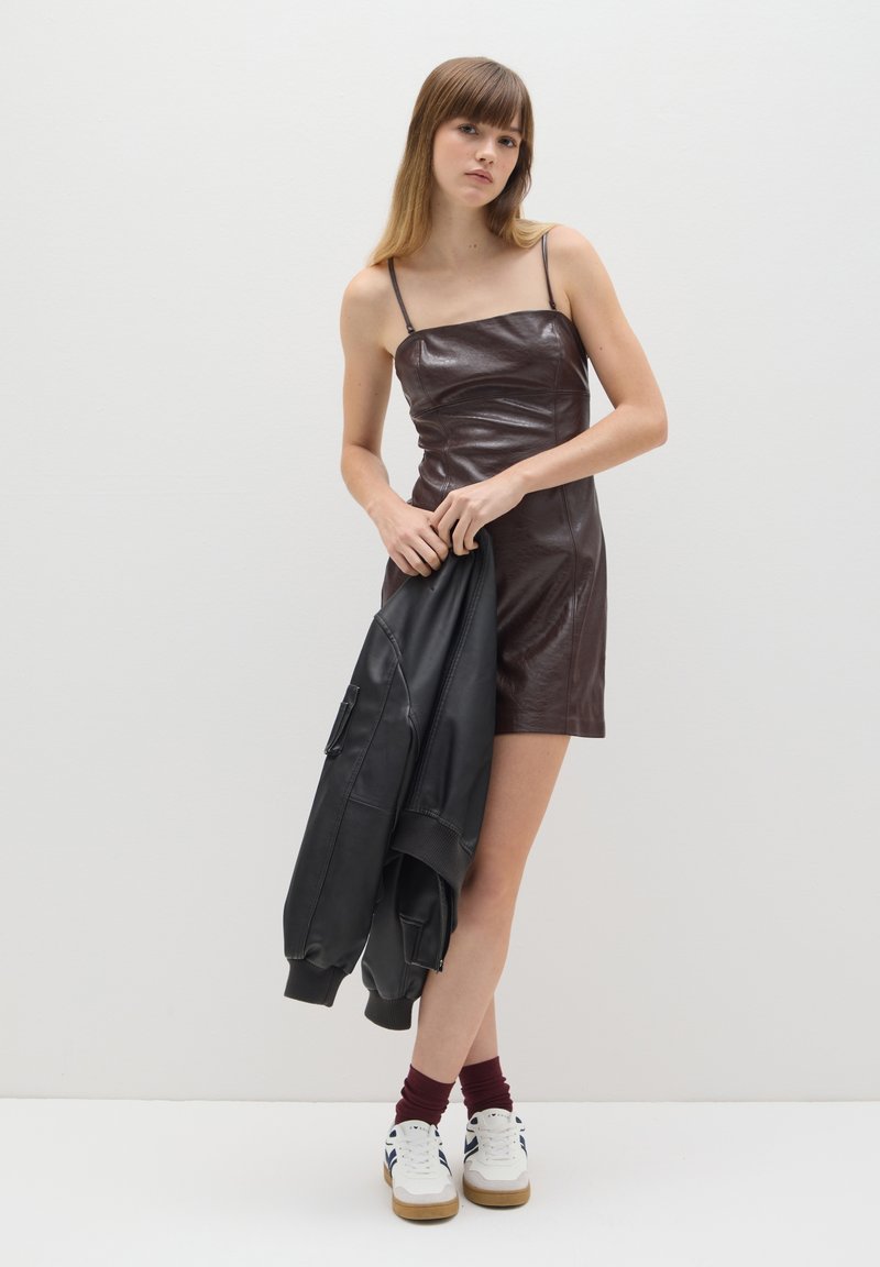 Vestido mini de cuero marrón con tirantes finos, diseño ajustado y textura brillante. La modelo sostiene una chaqueta bomber de cuero negra, viste calcetas burdeos y zapatillas.