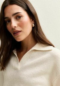 Kremowy sweter polo z dzianiny o strukturalnym wykończeniu i kołnierzykiem, w połączeniu ze złotymi kolczykami w kształcie kropli.
