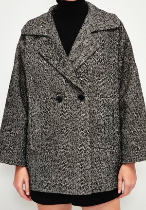 Femme portant un manteau croisé motif chevrons noir et blanc par-dessus un col roulé noir et un short noir.