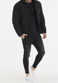 Černá péřová bunda s kapucí, zipovým zapínáním a žebrovanými manžetami, nošená na černém tričku a v kombinaci s černými Skinny džínami a bílými teniskami.