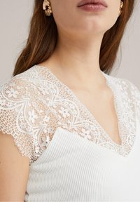 Vit topp med spetsärmar och blommönster, V-ringning och ribbat mönster på kroppen. Guldörhängen synliga.