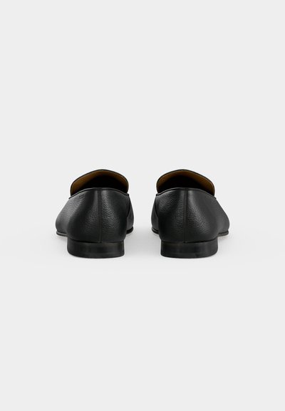 Paire de chaussures en cuir noir à enfiler, vue de dos sur un fond clair uni.