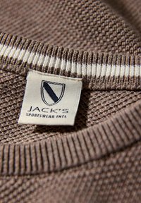 Brązowa tkanina o fakturze z prążkowanym kołnierzem, z metką z napisem „JACK'S SPORTSWEAR INTL” i trójkątnym logo w ciemnym niebieskim kolorze.