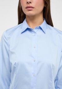 Camicia button-up azzurra chiara con colletto classico, caratterizzata da una texture liscia e sei bottoni sul davanti. Design semplice e pulito.