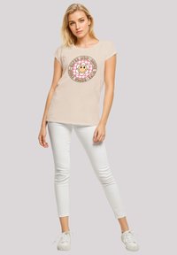 Beige cotton t-shirt featuring a circular graphic with a smiling lion and text, paired with white skinny jeans and white sneakers.