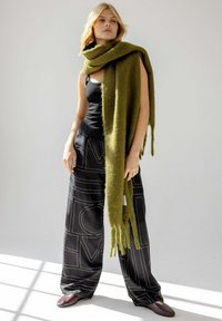 Écharpe en laine vert olive drapée librement, avec des bords frangés ; associée à un pantalon en satin noir arborant un motif audacieux, et des chaussures foncées.